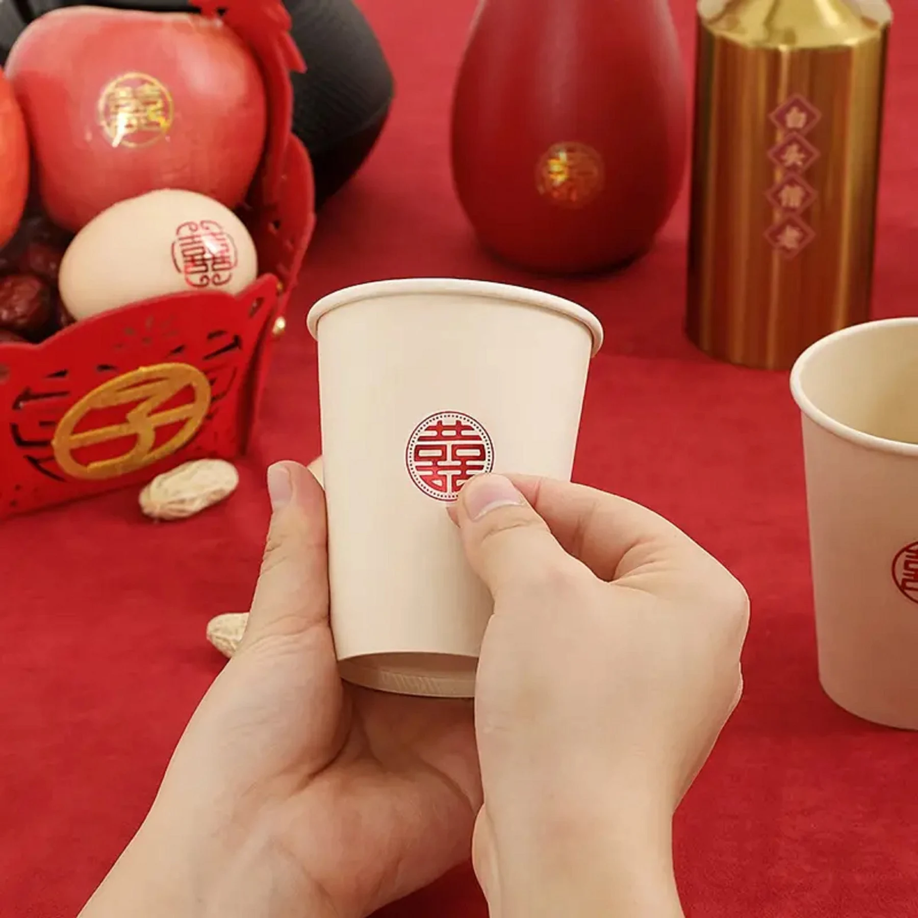 May include: A person holding a beige paper cup with a red double happiness symbol. The cup is part of a set of wedding decorations, including red apples, a red basket, and a red background.