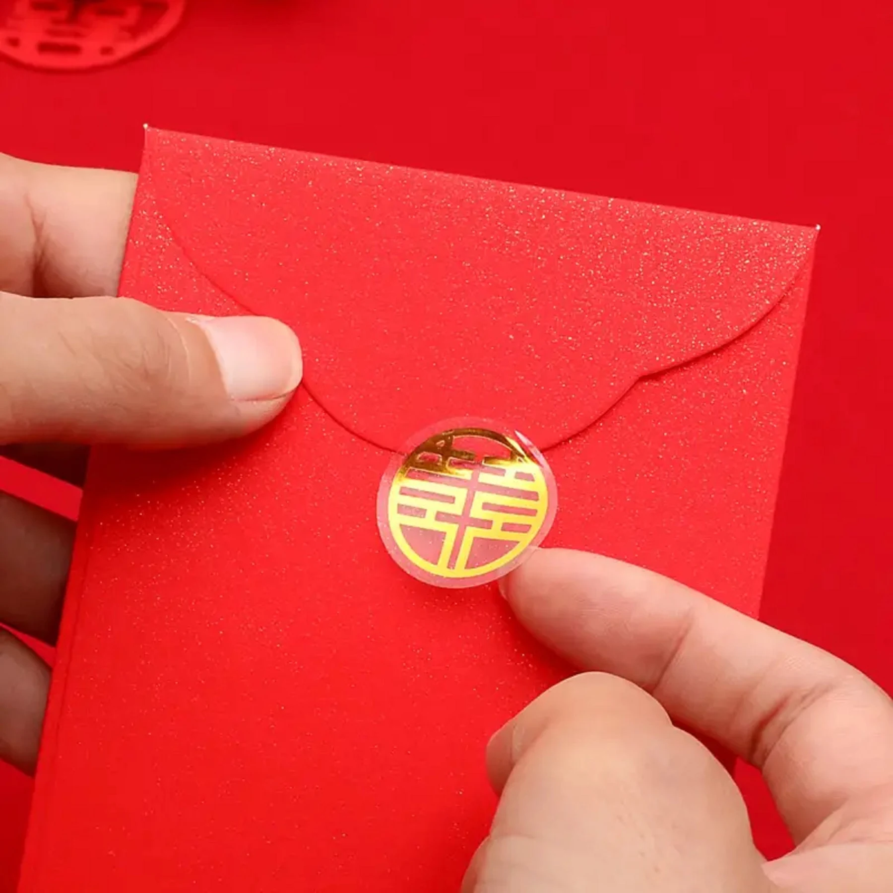 May include: A red envelope with a gold circular sticker featuring a Chinese double happiness symbol. The envelope is being held by two hands. The background is a solid red color.