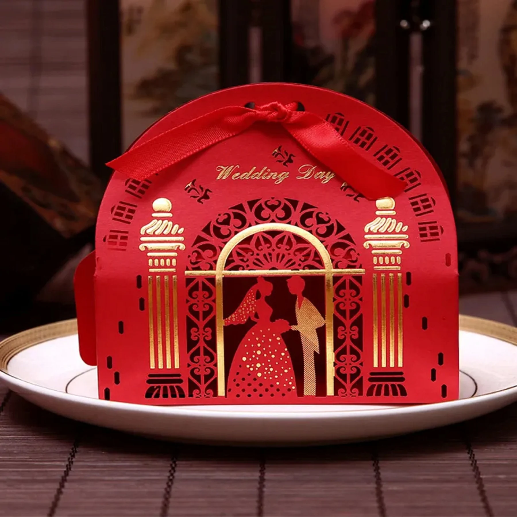May include: A red wedding favor box with a decorative cut-out design. The box features a silhouette of a bride and groom, gold accents, and the words "Wedding Day". A red ribbon is tied at the top.