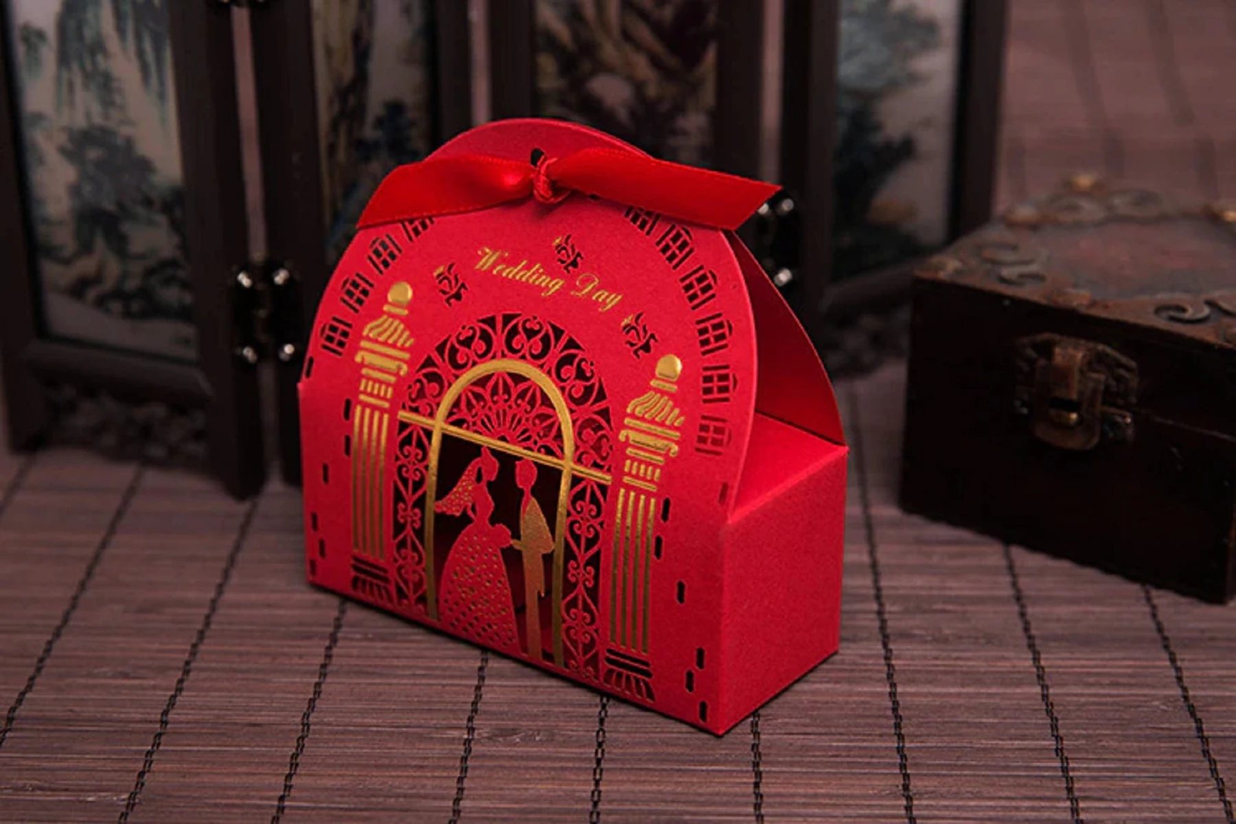 May include: Red wedding favor box with a decorative cutout design and a red ribbon. The box features gold accents and the words "Wedding Day". The design includes silhouettes of a bride and groom.