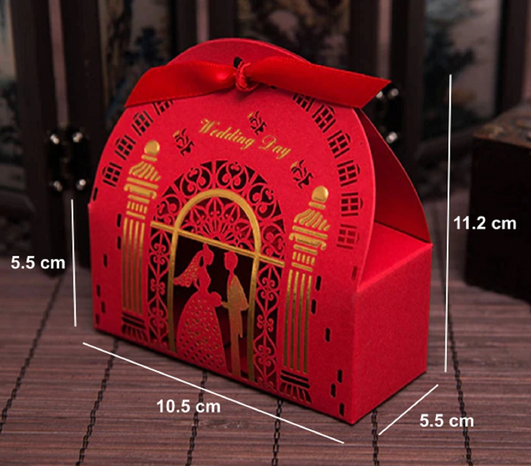 May include: Red wedding favor box with a handle and a red ribbon bow. The box is 11.2 cm tall, 10.5 cm wide, and 5.5 cm deep. It features a laser-cut design with a bride and groom silhouette and the words "Wedding Day".