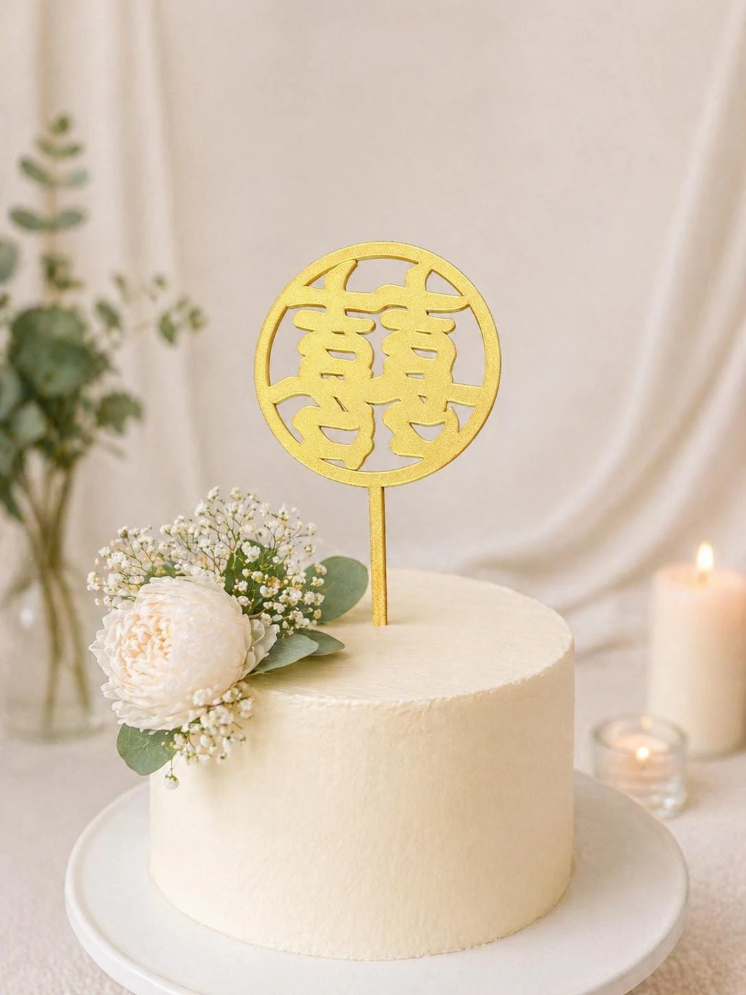 May include: A gold-colored circular cake topper featuring the Chinese double happiness character, mounted on a single stick. The topper is placed on a smooth, cylindrical white cake decorated with a cluster of white flowers and green leaves.