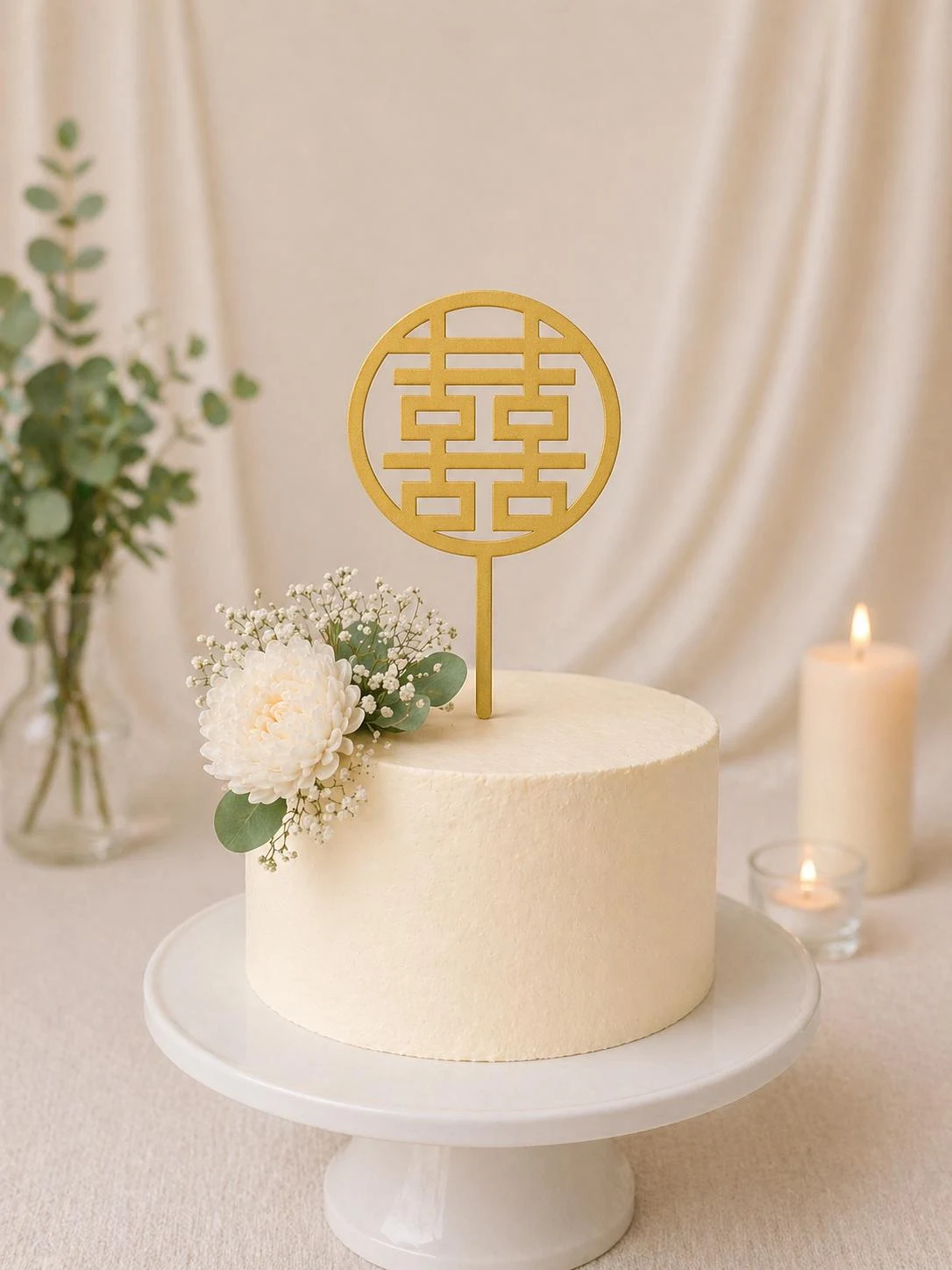 May include: A round gold cake topper featuring the Chinese double happiness symbol, positioned on top of a smooth, off-white frosted cake. The cake is decorated with a cluster of white flowers and green eucalyptus leaves on the side.