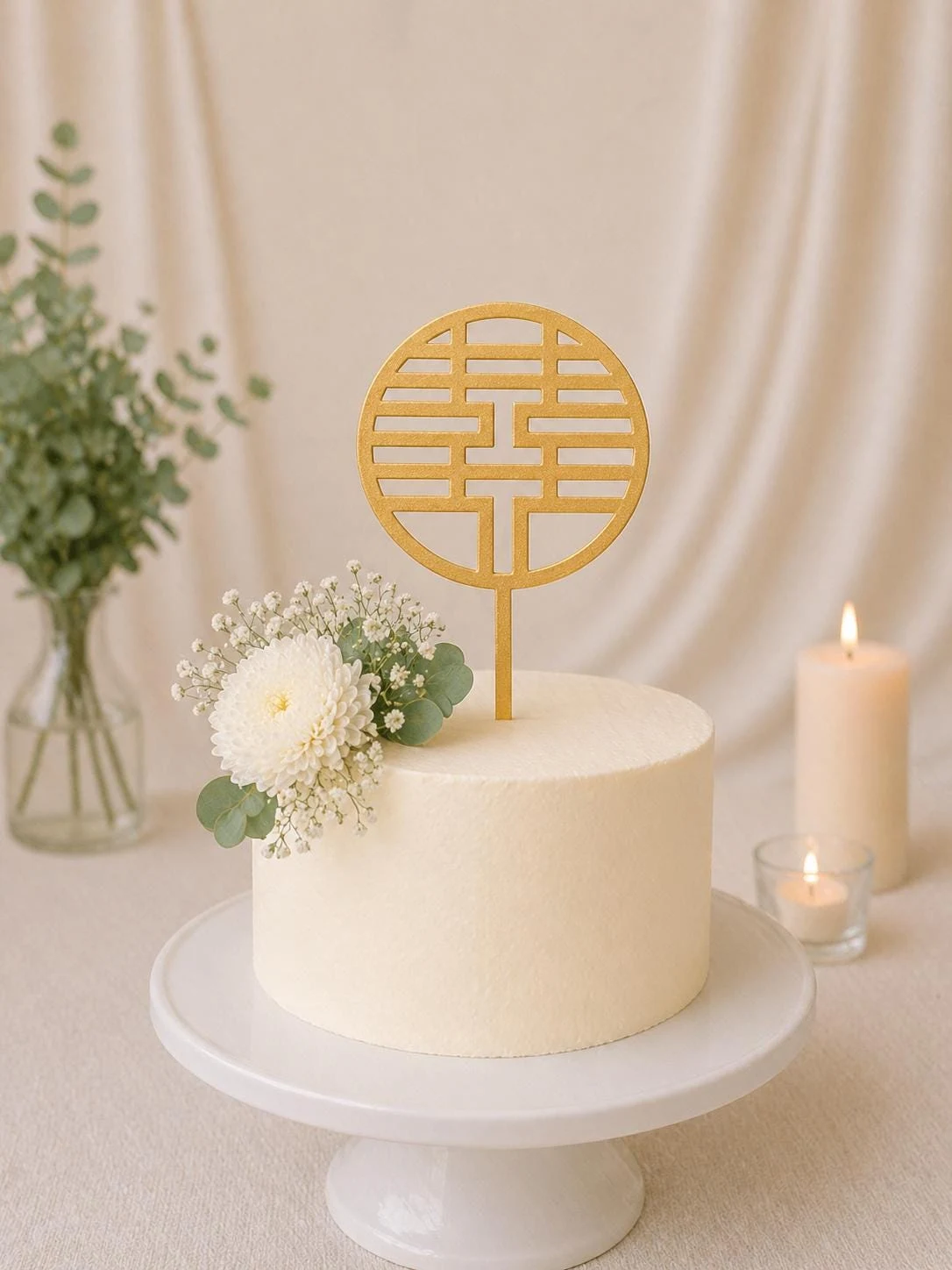 May include: A round, off-white cake on a white pedestal stand, decorated with a white chrysanthemum and baby's breath flowers. A gold-colored, circular double happiness symbol cake topper is inserted into the top center of the cake.