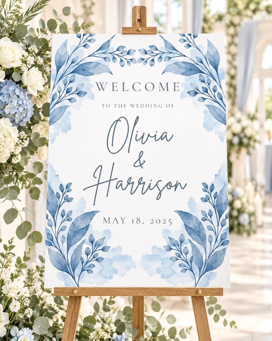 May include: A white wedding welcome sign on a wooden easel featuring blue watercolor botanical illustrations of leaves and branches. The text reads: WELCOME TO THE WEDDING OF Olivia & Harrison MAY 18, 2025 in dark blue serif and script fonts.
