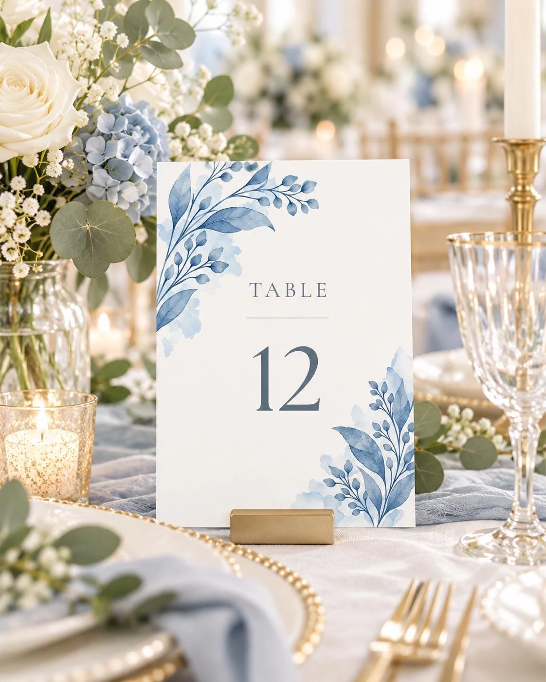 May include: A rectangular white table number card featuring a watercolor blue botanical design with leaves and berries in the top left and bottom right corners. The text reads TABLE 12 in a dark blue serif font, held by a gold metal stand.